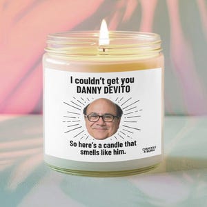 May include: A lit candle in a clear glass jar. The label features the text "I couldn't get you DANNY DEVITO" above an image of Danny DeVito and the text "So here's a candle that smells like him."