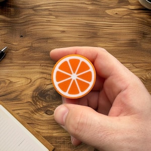 Könnte beinhalten: Ein runder orangener Schwamm mit weißem Rand und einem stilisierten Orangenscheiben-Design. Der Schwamm wird in einer Hand gehalten.