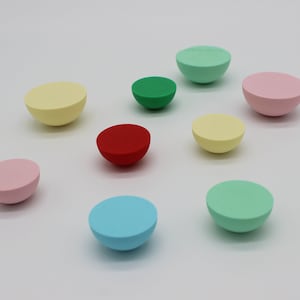 Puede incluir: Una colección de objetos coloridos en forma de media esfera en varios colores pastel, incluyendo amarillo, rosa, verde, rojo y azul. Los objetos están dispuestos sobre una superficie blanca, creando una exhibición lúdica y visualmente atractiva.