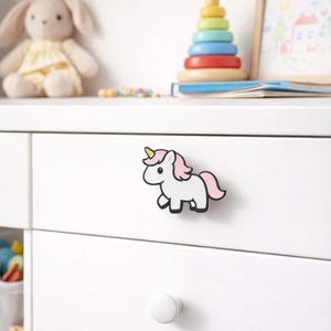 Może przedstawiać: Biały uchwyt do szuflady w kształcie jednorożca z różową grzywą i ogonem oraz żółtym rogiem. Jednorożec jest przymocowany do białej szuflady. Inne zabawki są widoczne w tle.
