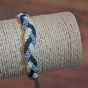 May include: Braided macrame bracelet in shades of blue and cream. The bracelet is displayed on a beige, textured spool. The macrame design features a woven pattern, showcasing the intricate craftsmanship of the handmade accessory.