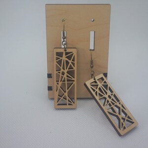 May include: Wooden geometric earrings with a rectangular shape and intricate cut-out design. The earrings are light brown and hang from gold-colored hooks. They are displayed on a wooden stand.