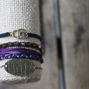 May include: A set of bracelets featuring a variety of textures and colours, including black, purple, and brown. The bracelets are stacked on a burlap-covered display stand. One bracelet has a gold lock charm.