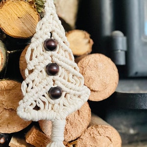 Może przedstawiać: Biała ozdoba choinkowa z makramu z brązową drewnianą koralikiem na górze i dwoma innymi koralikami w środku. Ozdoba jest zawieszona na kawałku drewna i otoczona stosem kłód.