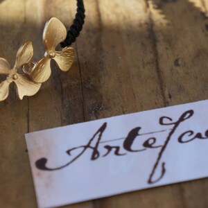 Könnte beinhalten: Ein goldenes Armband mit einem schwarzen Band und drei Blumenanhängern. Das Armband liegt auf einer Holzoberfläche mit einer weißen Karte, auf der "Artefweet" steht.