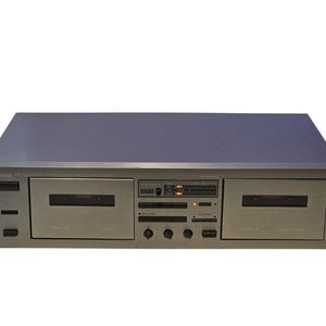 May include: A silver Yamaha dual cassette deck featuring two cassette compartments, a central control panel with various buttons, knobs, and a digital display. The unit has a rectangular metallic chassis with black accents and clear labels.