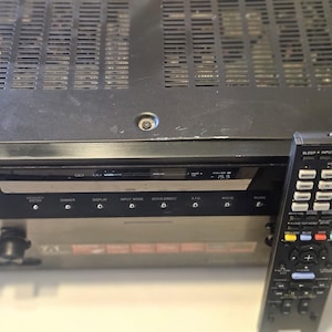 May include: A black audio receiver with a remote control. The receiver has a display screen and several buttons labeled "Memory Enter", "Dimmer", "Display", "Input Mode", and "2CH/Direct". The remote control has buttons for various functions.