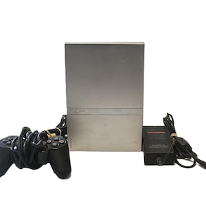 May include: A silver Sony PlayStation 2 game console standing vertically, accompanied by a black wired game controller and a black power adapter with a cord. The console features the PS2 logo on the front and the Sony brand name vertically.