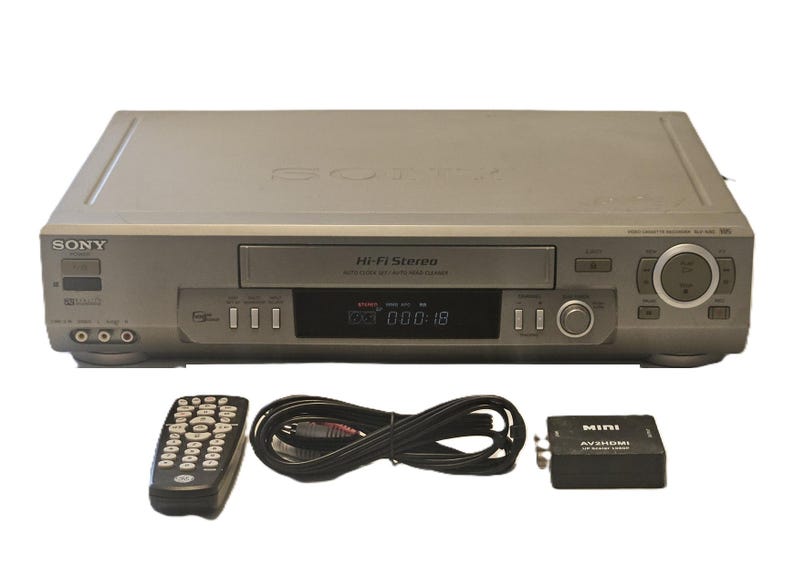 May include: A silver Sony Hi-Fi Stereo VHS player with a digital display showing 000:18. Included are a black remote control, a coiled black RCA cable, and a black Mini AV2HDMI converter box with the text MINI AV2HDMI 1080P printed on top.
