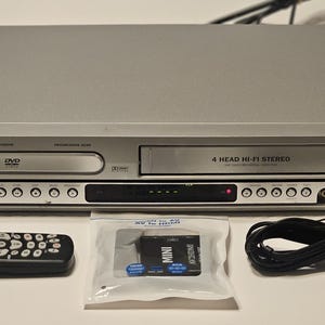 May include: A silver Magnavox DVD/VCR combo player with a remote, AV cables, and a Mini AV to HDMI adapter. The player has a DVD tray and a VHS tape slot. The remote is black with white buttons.