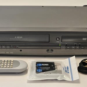 May include: A gray Magnavox DVD/VCR combo player with a remote, cables, and a Mini V2HDMI adapter. The VCR has "4 HEAD" printed on it. The DVD player has a "DVD" logo. The remote is gray with many buttons.