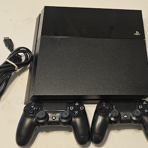 May include: A black PlayStation 4 game console with a matte and glossy finish, featuring the PlayStation logo. Included are two black wireless game controllers with directional pads, analog sticks, and action buttons, plus a black HDMI cable.