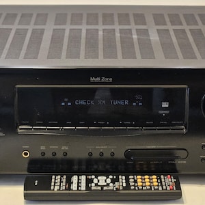 May include: Black Denon AVR-2300CI receiver with a remote control. The receiver has a display that reads "CHECK XM TUNER". The receiver has multiple buttons and knobs, including a large volume knob on the right side.