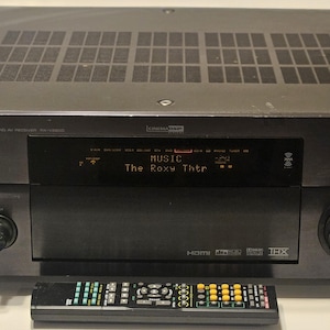 May include: A black Yamaha receiver with a display showing "MUSIC The Roxy Thtr". The receiver has multiple buttons and a large volume knob. A remote control is in front of the receiver.