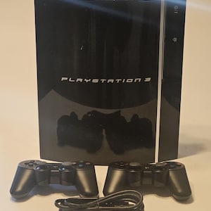 May include: A black PlayStation 3 console with two matching black controllers and a black cable. The console has the words "PLAYSTATION 3" printed on the front. The controllers are positioned in front of the console.