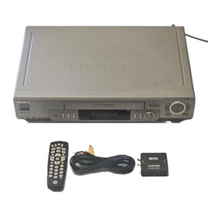 May include: A silver Sony VCR player with Hi-Fi Stereo branding, featuring a front-loading tape slot and digital display. Included are a black remote control, a coiled black RCA cable, and a small black Mini AV2HDMI upscaler converter box.