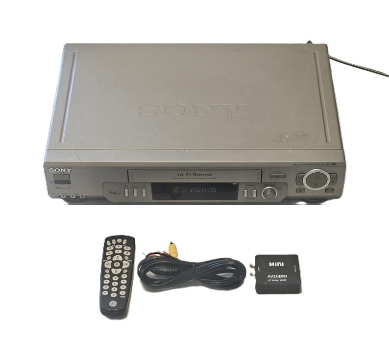 May include: A silver Sony VCR player with Hi-Fi Stereo branding, featuring a front-loading tape slot and digital display. Included are a black remote control, a coiled black RCA cable, and a small black Mini AV2HDMI upscaler converter box.