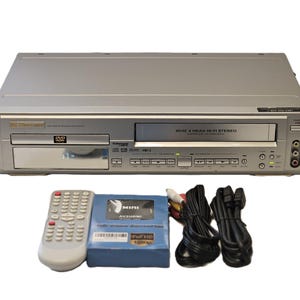 May include: Silver DVD/VCR combo player with a remote, a blue AV/HDMI adapter, and various cables. The player has a DVD tray and a VCR slot. The remote is white with many buttons. The adapter is labelled "MINI AV/HDMI".
