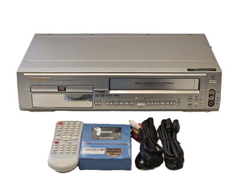 May include: Silver DVD/VCR combo player with a remote, a blue AV/HDMI adapter, and various cables. The player has a DVD tray and a VCR slot. The remote is white with many buttons. The adapter is labelled "MINI AV/HDMI".