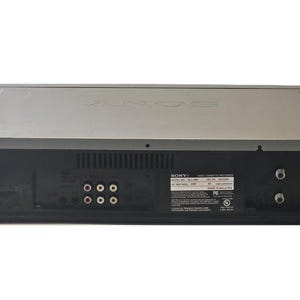 May include: A silver and black Sony video cassette recorder viewed from the back panel. The device features multiple RCA input and output ports, two coaxial connectors, a power cord, and a rectangular label with technical specifications and text.