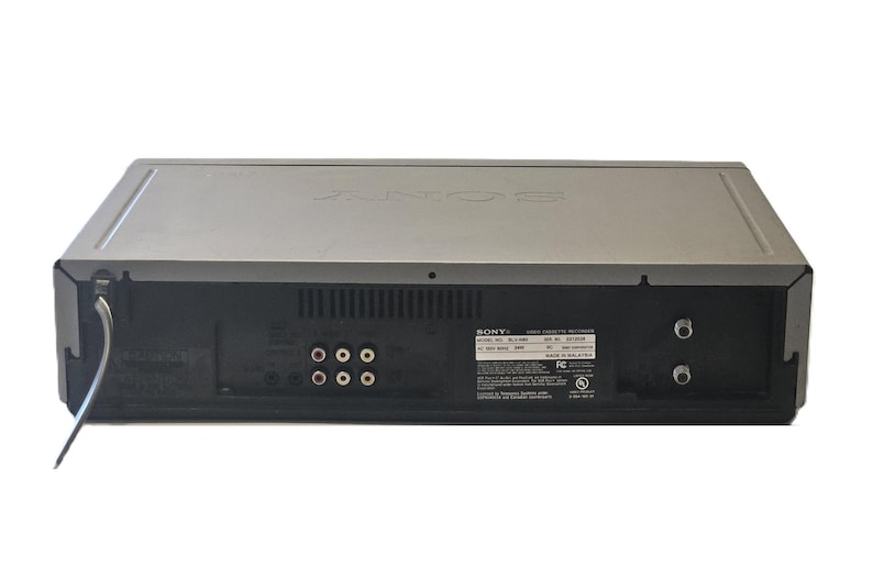 May include: A silver and black Sony video cassette recorder viewed from the back panel. The device features multiple RCA input and output ports, two coaxial connectors, a power cord, and a rectangular label with technical specifications and text.