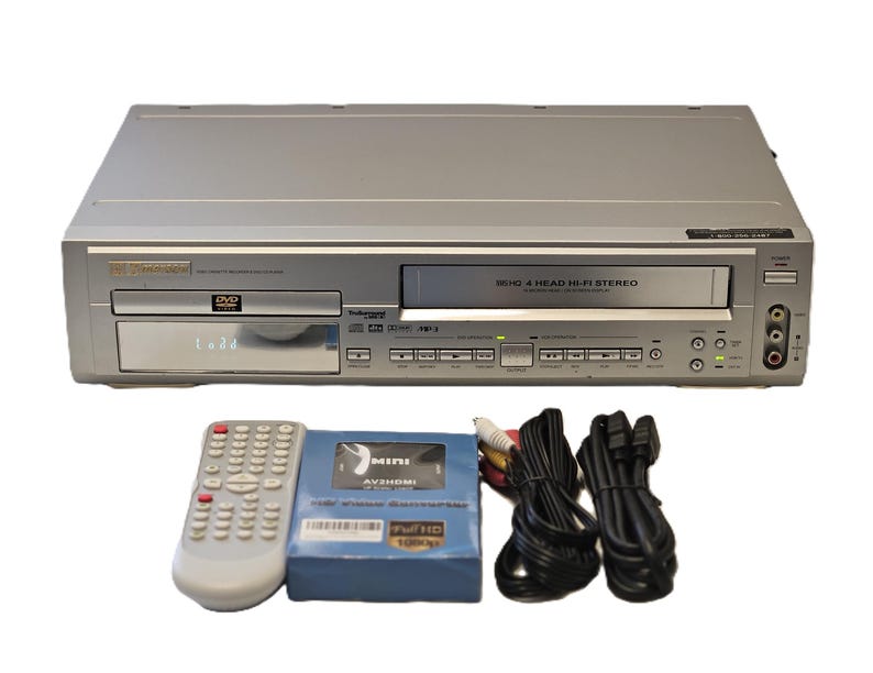 May include: Silver DVD/VCR combo player with a remote, a blue AV/HDMI video converter, and black cables. The player has a DVD tray and a VHS tape slot. The remote is white with many buttons. The converter is in a blue box.