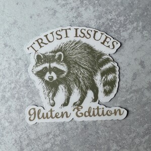 May include: A sticker featuring a detailed illustration of a raccoon in shades of gray and brown. The text "TRUST ISSUES" arches above the raccoon, and "Gluten Edition" is below. The sticker has a white border.