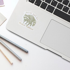 May include: A white sticker with a raccoon illustration and the text "TRUST ISSUES Gluten Edition" is affixed to a laptop. Two pencils are on the white surface. The raccoon is in a light green color.