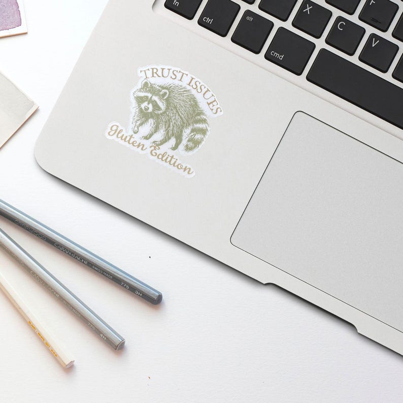 May include: A white sticker with a raccoon illustration and the text "TRUST ISSUES Gluten Edition" is affixed to a laptop. Two pencils are on the white surface. The raccoon is in a light green color.