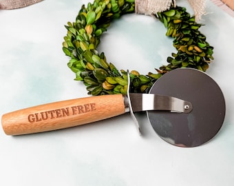 May include: A wooden pizza cutter with a stainless steel blade. The handle is engraved with the words "Gluten Free".
