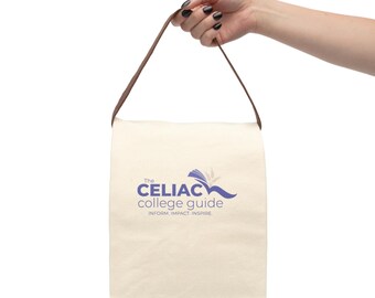 May include: Cream-colored tote bag with a brown handle, held by a hand. The bag features the text "The CELIAC college guide" in blue, with a stylized logo. The bag is designed for carrying items.