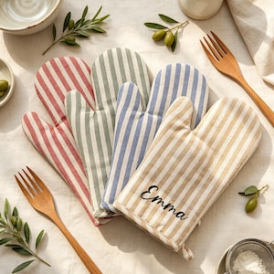 May include: Four oven mitts featuring vertical stripes in red, sage green, blue, and beige. The beige mitt is embroidered with the name Emma in black script. The mitts are arranged in a fan shape on a light-colored fabric surface with wooden forks.