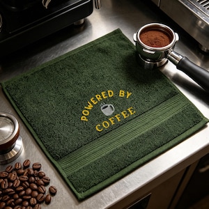 Puede incluir: Una toalla cuadrada de algodón verde oscuro con el texto amarillo bordado "POWERED BY COFFEE" arqueado sobre un pequeño icono de taza de café gris. La toalla tiene una superficie de rizo texturizada con una banda horizontal lisa en la base.