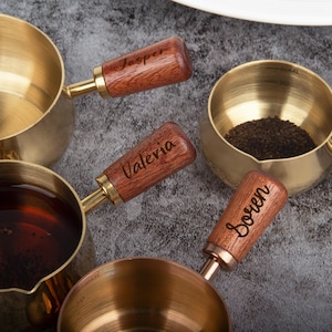 May include: Several brass measuring cups with wooden handles. The handles are engraved with the names "Jasper", "Valeria", and "Soren". One cup contains tea leaves, and another contains a dark liquid. The cups have measurement markings.