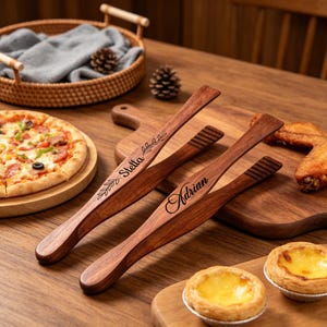 May include: Three wooden serving tongs with personalized names "Stella" and "Adrian" engraved on them. The tongs are dark brown and have a sleek design. The image also shows pizza, pastries, and other food items.