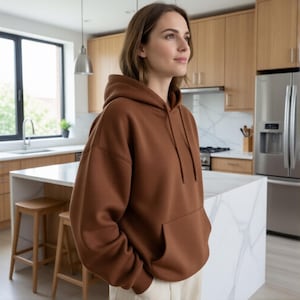 May include: A brown hooded sweatshirt with a front pocket and drawstring. The sweatshirt is made of a soft material and is worn by a person. The background shows a kitchen with wooden cabinets and a marble countertop.