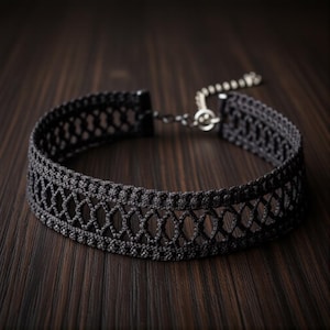 Op de afbeelding: Een zwarte choker ketting met een open ontwerp. De ketting is gemaakt van een geweven materiaal en heeft een delicaat patroon. De choker heeft een zilverkleurige sluiting en ketting voor verstelbare maatvoering. De achtergrond is een donker houten oppervlak.
