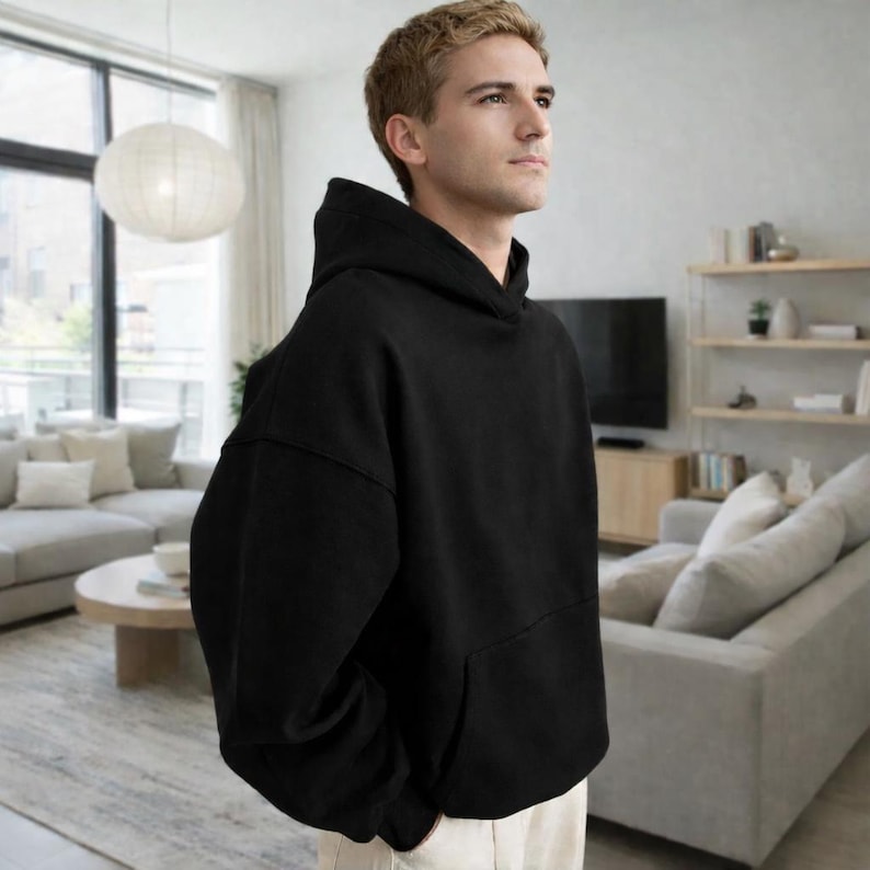 May include: A black hoodie with a front pocket and a hood. The hoodie is made of a thick, soft material. The person is wearing light-colored pants.