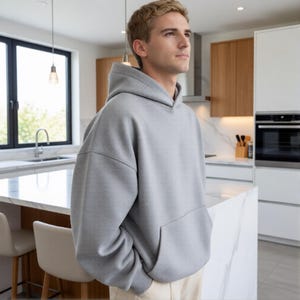 May include: A light gray hoodie with a kangaroo pocket. The hoodie is made of a soft, thick material and has a drawstring hood. The person is wearing beige pants. The background is a modern kitchen with white cabinets and a marble countertop.