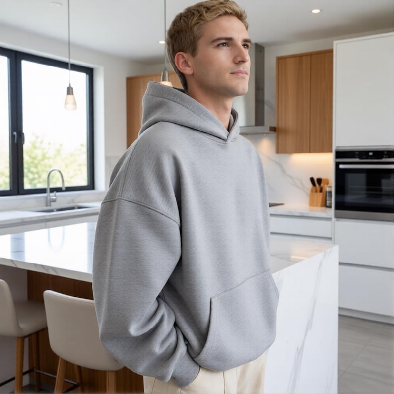 May include: A light gray hoodie with a kangaroo pocket. The hoodie is made of a soft material and has a relaxed fit. The person is wearing beige pants. The background is a modern kitchen with white cabinets and a marble countertop.