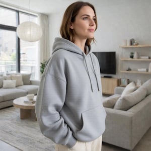 May include: A light gray hoodie with a front pocket and drawstring hood. The sweatshirt is made of a soft material and is a casual style. The person is wearing the hoodie in a living room setting.