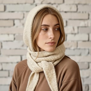 May include: A cream-colored knitted hood and scarf set. The hood frames the face, and the scarf drapes down the front. The knitwear has a textured appearance, and the color is a soft, neutral tone. The background is a light-colored brick wall.
