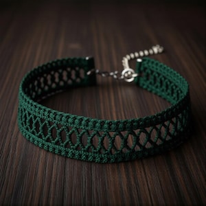 May include: A dark green choker necklace with an openwork design. The necklace is made of a woven material and features a silver-colored chain and clasp. The choker is displayed on a dark wooden surface.