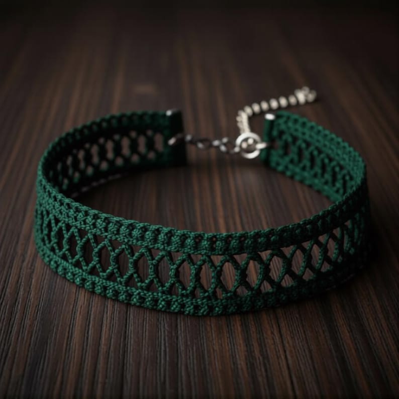 May include: A dark green choker necklace with an openwork design. The necklace is made of a woven material and features a silver-colored chain and clasp. The choker is displayed on a dark wooden surface.