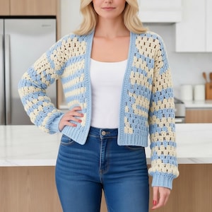 Könnte beinhalten: Eine hellblaue und cremefarbene Häkeljacke mit langen Ärmeln. Der offene Pullover hat ein Granny-Square-Muster. Die Ärmel und der untere Saum haben ein Rippendesign. Die Jacke wird über einem weißen Oberteil und einer blauen Jeans getragen.