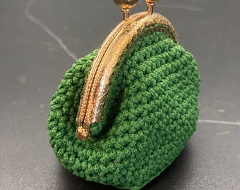 May include: A small, green crocheted coin purse with a gold-coloured clasp. The clasp features an ornate floral design and two small, green gemstone accents. The purse is closed.