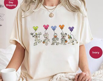 May include: A white t-shirt with a print of Mickey Mouse and friends dressed as mummies holding balloons. The balloons are different colors, including yellow, blue, green, orange, and purple. The shirt is a light ivory color.