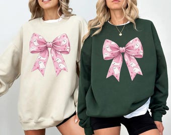 May include: Two sweatshirts, one cream and one dark green, each featuring a large pink bow graphic. The bow design includes white ghost illustrations and small pink hearts. The sweatshirts are long-sleeved.