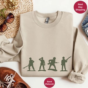 Puede incluir: Una sudadera de color arena con un gr&aacute;fico de cinco soldados de juguete en fila. Los soldados son verdes y sostienen varias armas. El texto "Arena (Natural)" est&aacute; impreso debajo del gr&aacute;fico.