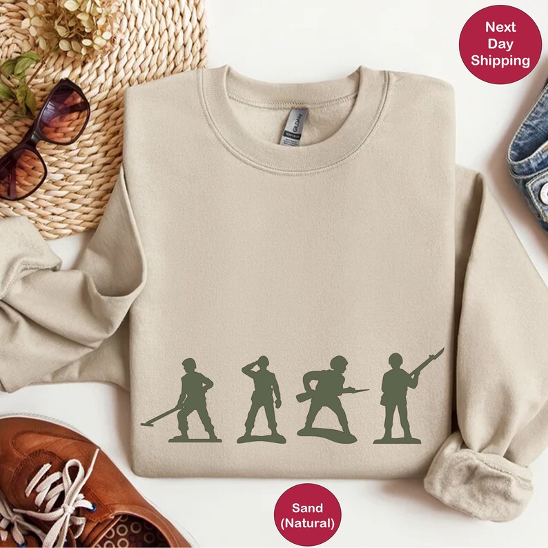 Puede incluir: Una sudadera de color arena con un gr&aacute;fico de cinco soldados de juguete en fila. Los soldados son verdes y sostienen varias armas. El texto "Arena (Natural)" est&aacute; impreso debajo del gr&aacute;fico.